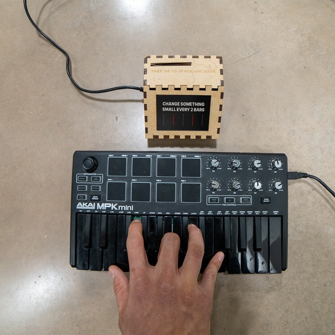 Hand plays an Akai MPK Mini below a laser-cut wooden coach box prompting: change something small every 2 bars
