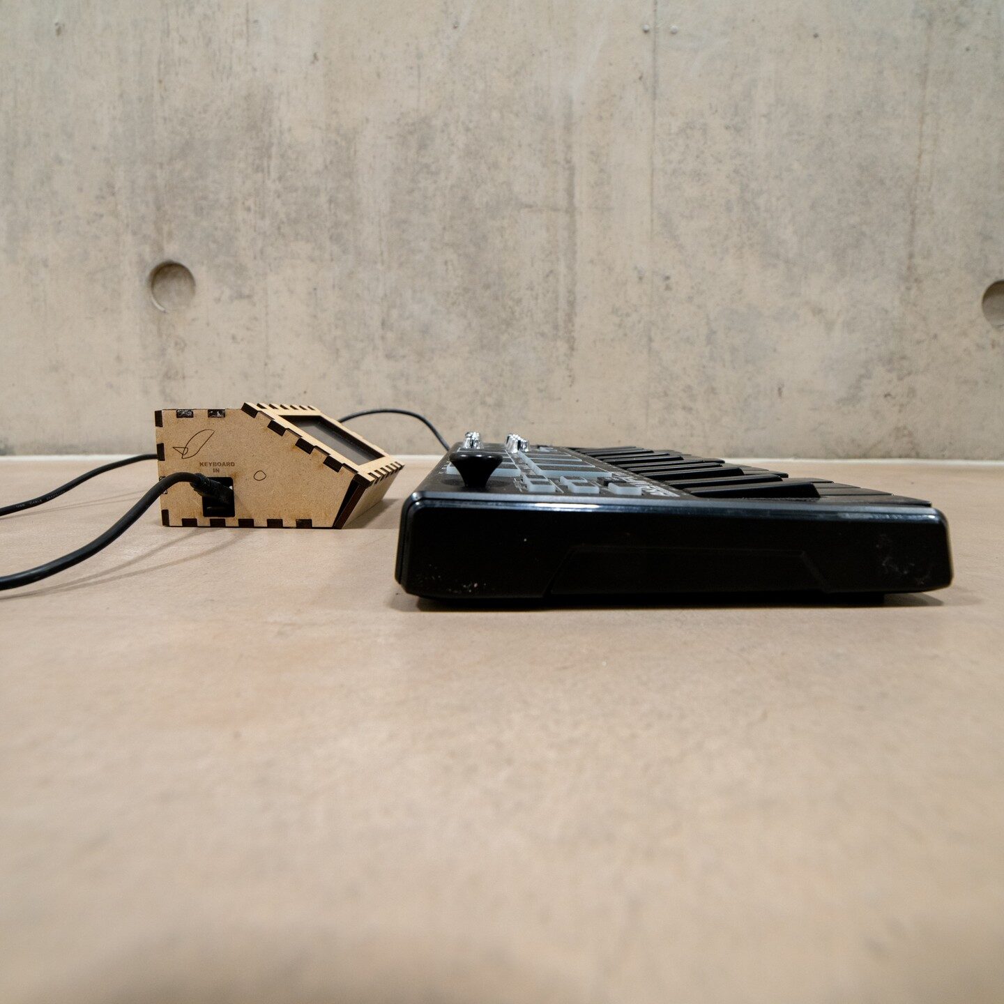 Side profile of the plywood improv-coach box cabled to an Akai MPK Mini against a concrete wall