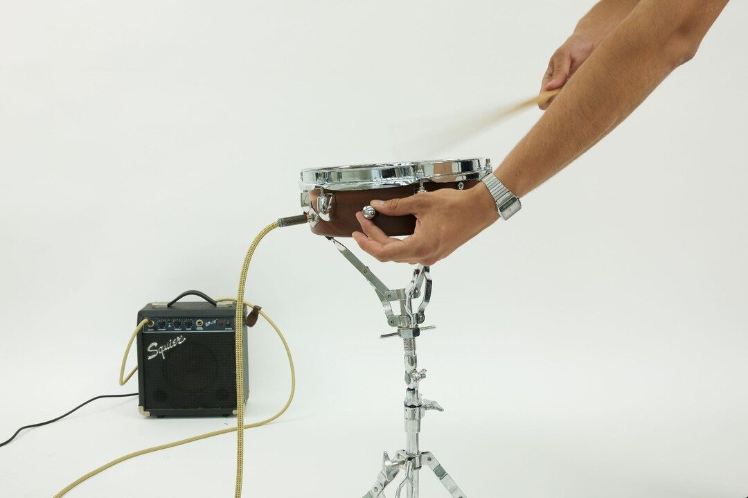 Hand adjusting the drum's onboard volume knob while a drumstick hovers, small amp in the background
