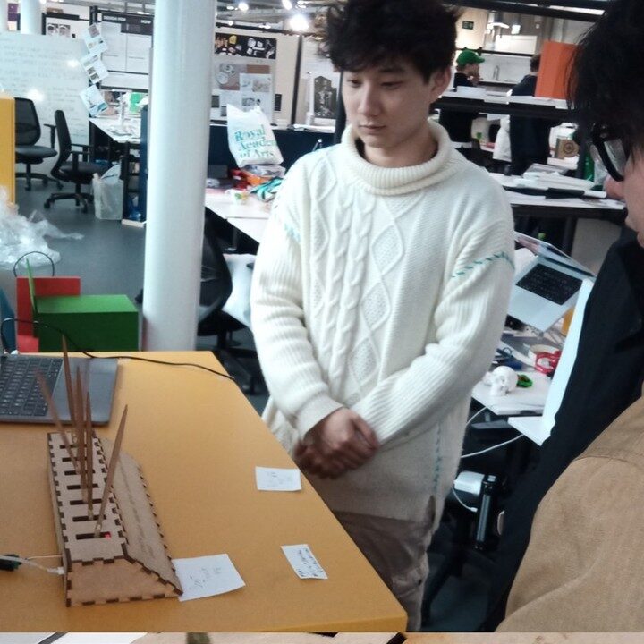 Maker presents the SEXTET prototype on a yellow desk to a visitor in a busy design studio