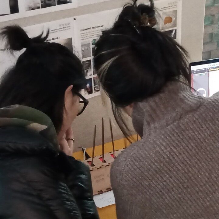 Two visitors lean in close over the SEXTET prototype beneath pinned project boards